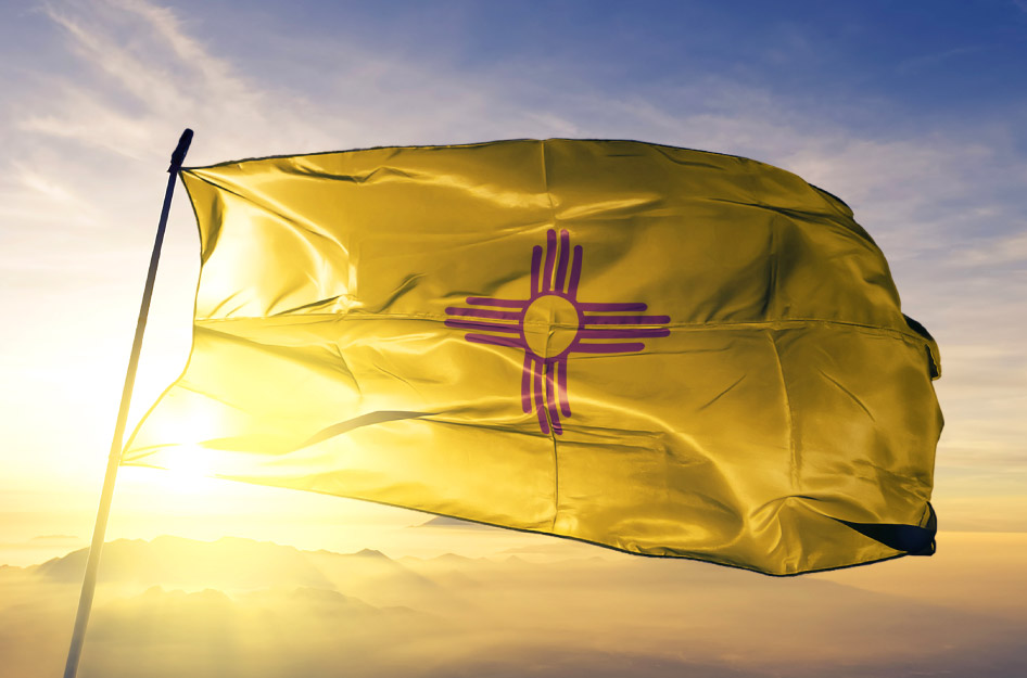 The state flag of New Mexico flies in the wind at the top of a flag pole as the sun rises behind a range of mountains in the distance. The flag features a yellow field with a red symbol in the center, representing the Sun.