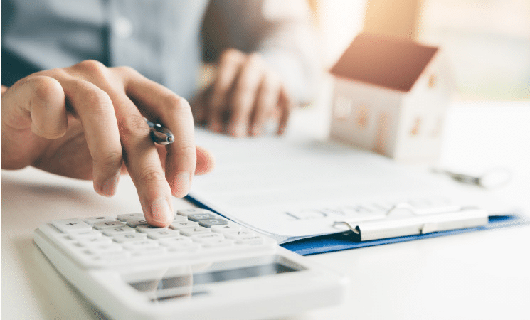 A close-up of a man using a calculator to calculate his home-buying budget. He is holding a pen and there is a clipboard with papers and a small model of a house on the table.