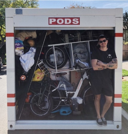 A man in a black top, black shorts, sunglasses and tattoos poses in front of a fully-loaded PODS moving and storage container.
