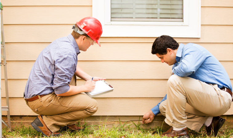 A home inspector and his assistant examine the foundation of a residential home. The inspector is pointing out a crack in the concrete beneath the siding. 
