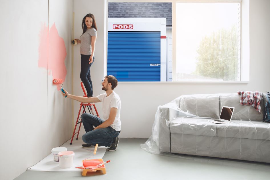 A young couple completes a fast home project by painting the walls of their new home. Their closed PODS container can be seen through the window.