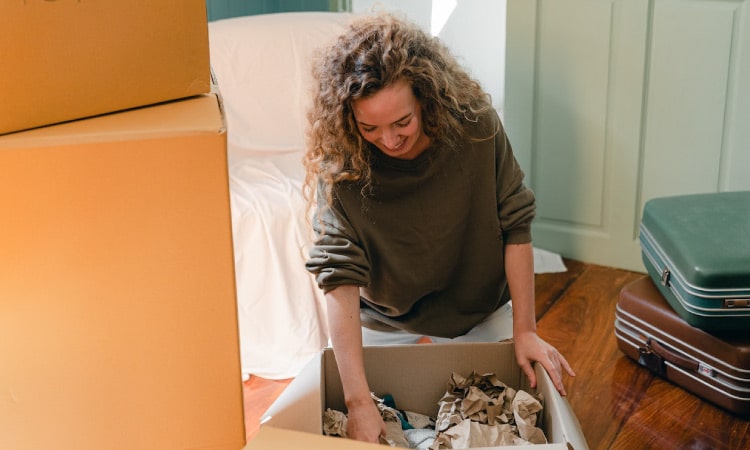 A woman with long, curly hair is kneeling on the floor while she unpacks a moving box. There is a stack of moving boxes beside her and a pair of suitcases on the floor behind her.