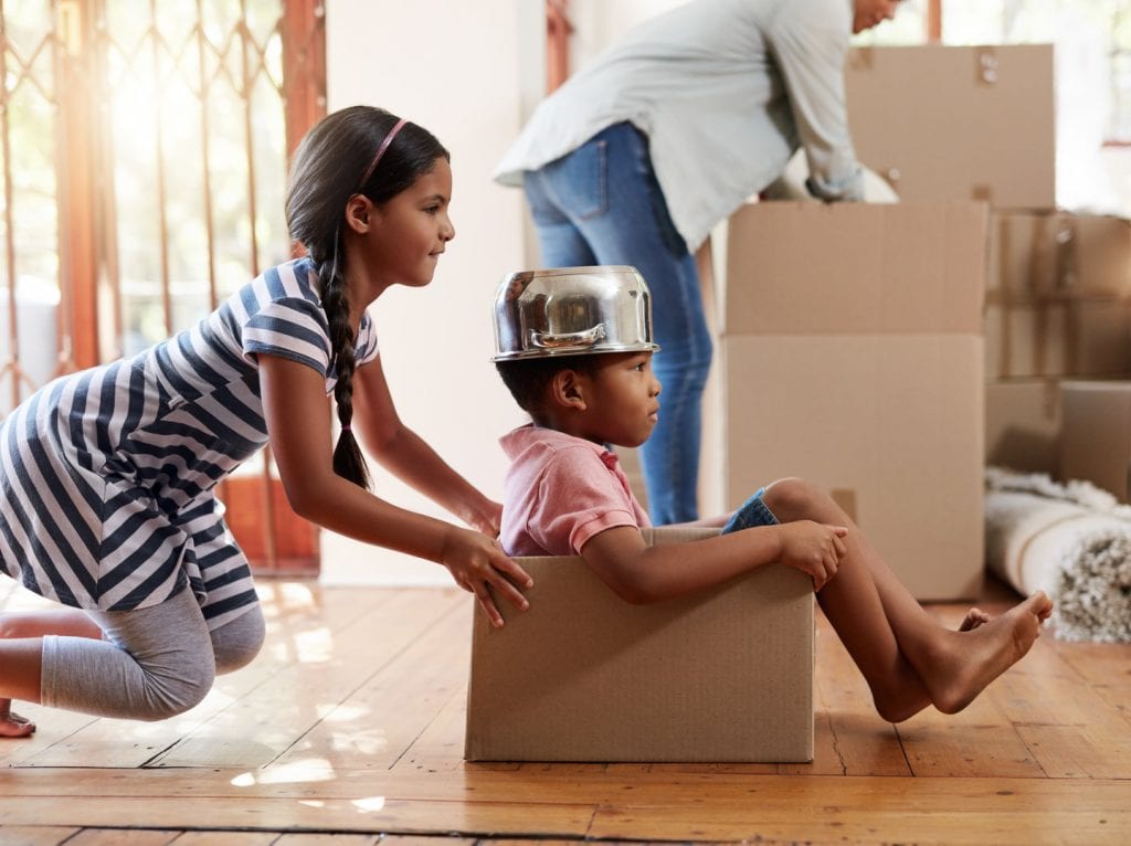 Two young children are playing with an empty moving box as their mother is packing another larger box in the background.