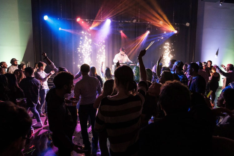 A DJ putting on a show at The Electric Belle at Stovehouse in Huntsville, Alabama. The dark room is filled with people who are dancing to the music. Lights on the stage send streams of orange and blue across the dance hall. 