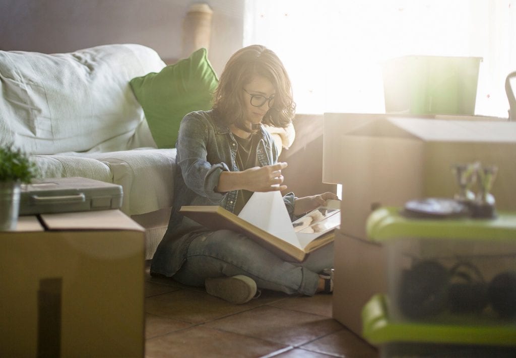 A college student sits in their apartment as the sun shines through. The woman is surrounded by cardboard boxes as she looks through a photo album.