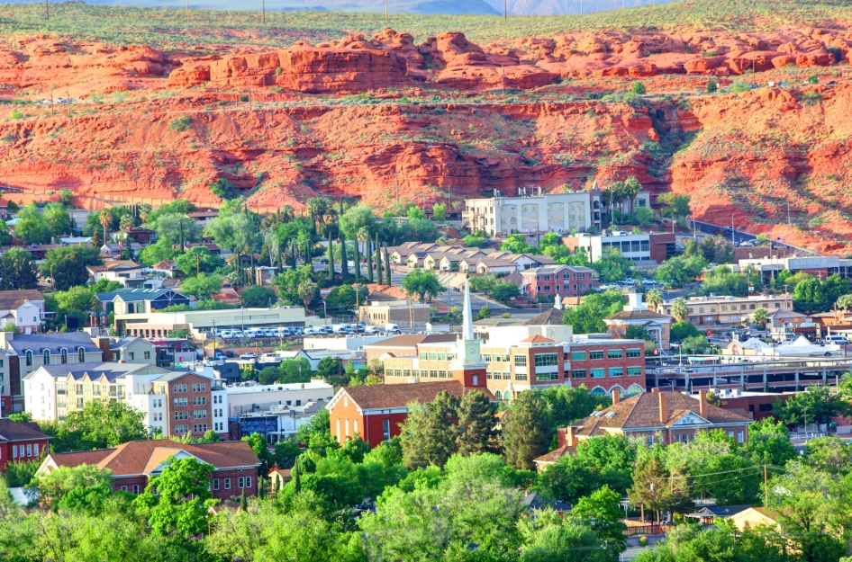 A scenic view of city buildings nestled among lush green trees with vibrant red cliffs visible in the background — a common view for those living in St. George, Utah.
