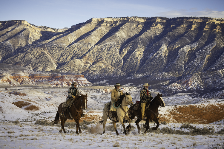 Two men and a woman are riding on horseback across a snowy Wyoming ranch with stunning mountains in the background.