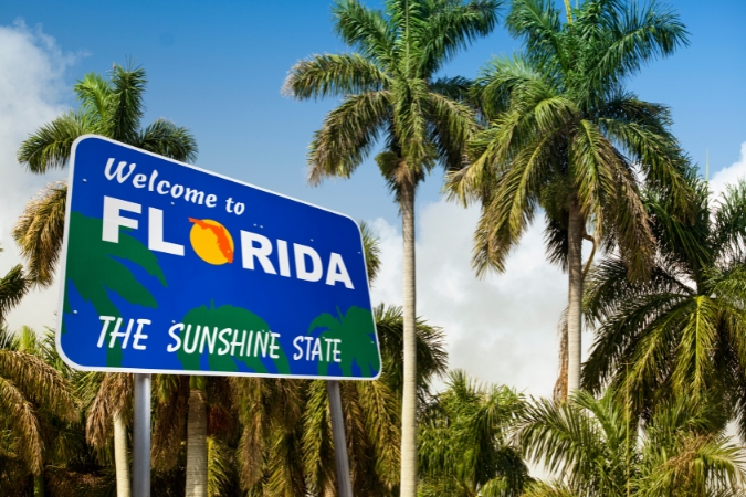 A large and colorful “Welcome to Florida” sign along a highway, surrounded by palm trees and cloudy skies, greeting drivers as they move down South to enter the Sunshine State.