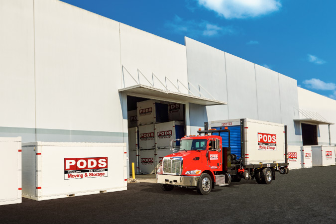 Exterior view of a PODS Storage Center with a truck and several containers parked just outside and stacked containers visible inside.