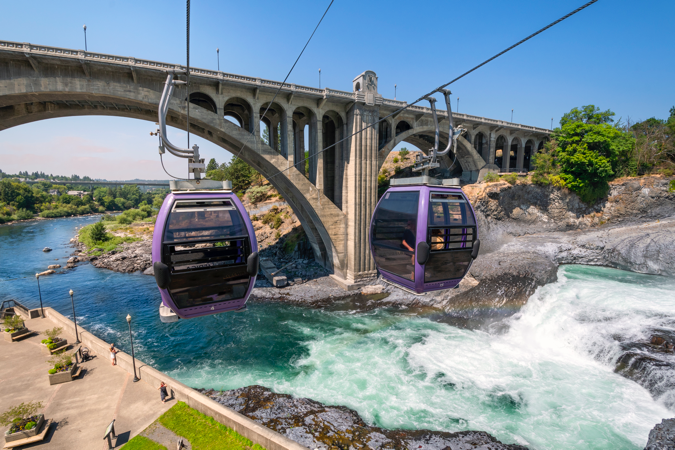 Two purple gondolas fly over the Spokane River and fall near Riverfront Park on a sunny day with clear skies.