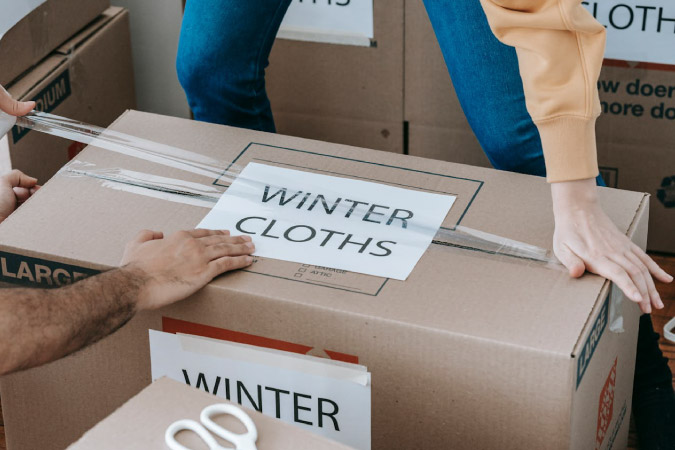 Close up of two pairs of hands taping up a moving box labeled “WINTER CLOTHES” as they prepare for moving out of state.
