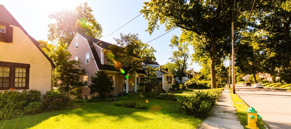 Street-level view of a neighborhood in New Rochelle, New York. The homes are two stories and feature green lawns on a tree-lined street.