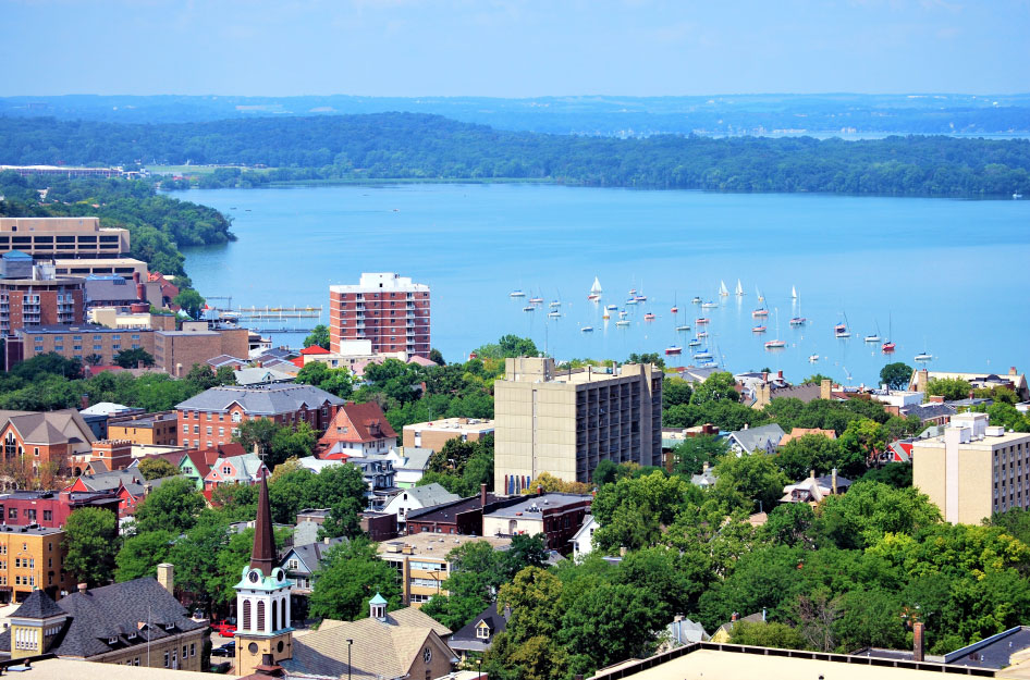 Aerial view of Downtown Madison, Wisconsin, and the surrounding forests and lakes. There are many sailboats out on the water, and the mature trees in the downtown area are lush with green leaves.