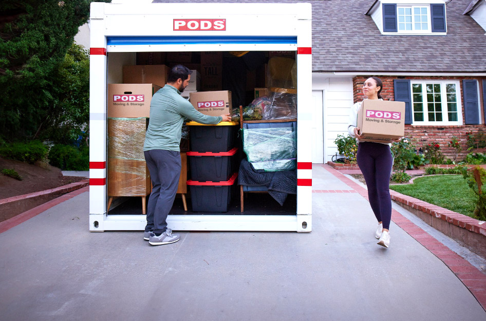 A couple is loading moving boxes into a PODS portable storage container in their driveway.