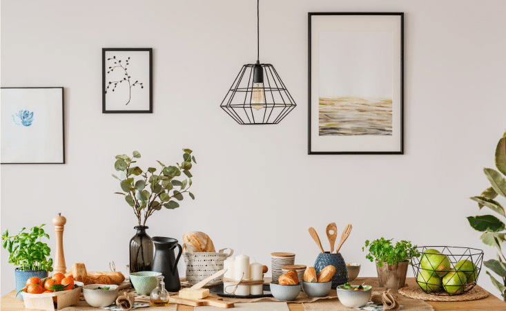 A white wall with three pieces of nature-themed art hanging on it. There’s an abstract waterscape, a simple painting of a berry branch, and a watercolor of a single blue flower. The butcher block island in front of the wall is filled with house plants, wooden utensils and baskets of fresh fruits and vegetables.