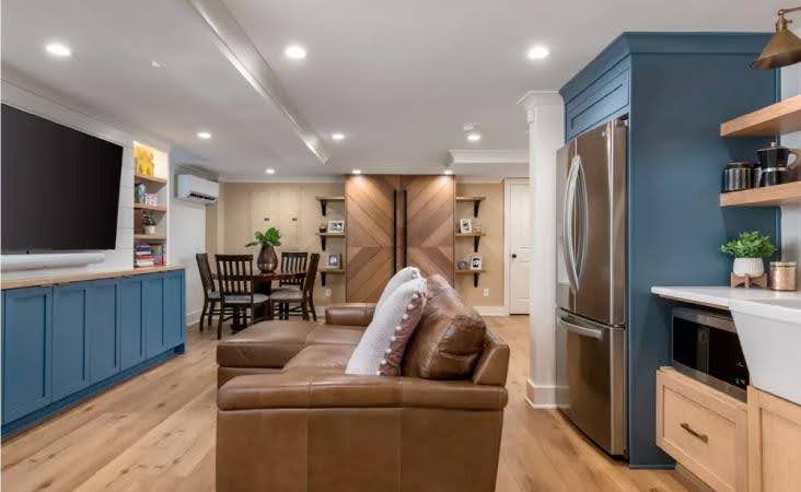 A basement that has been remodeled into a separate living suite. The walls are painted white, and the built-in cabinets are painted in a blue accent color. There is recessed overhead lighting, wood flooring, and a kitchenette with a full-sized fridge set into the wall.