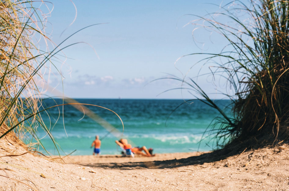 The view through a sandy dune path to a mature couple relaxing on a Florida beach. The man is fishing, and the woman is sunning nearby.