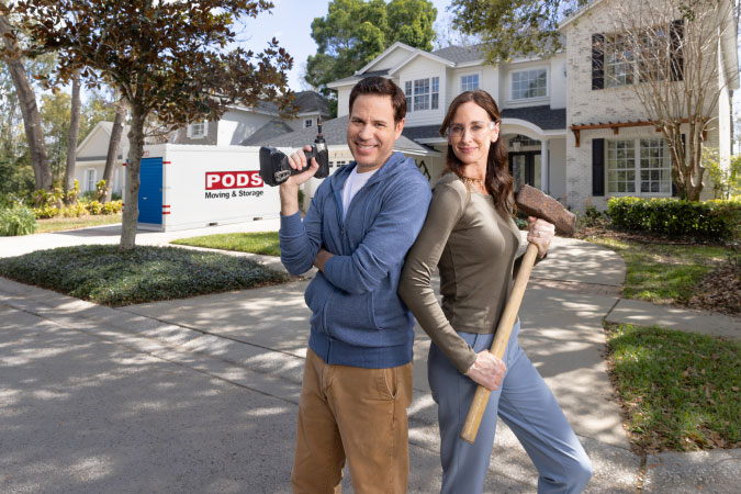 A couple poses with a drill and a sledgehammer in front of their home and a PODS portable storage container parked in their driveway.