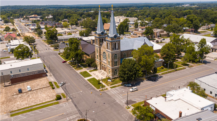 Aerial view of Cullman, Alabama, on a sunny summer day. The streets are quiet with very few cars, and  lush woods fill the gaps between many buildings and make up the space beyond the city.