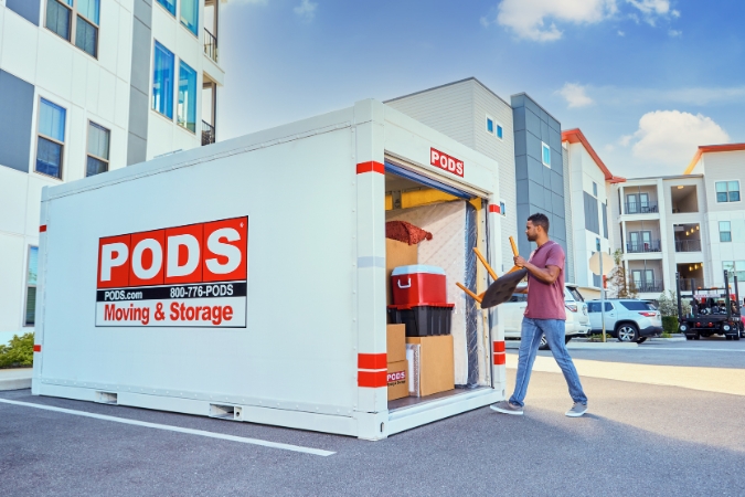 A man is in the process of moving out of an apartment, loading furniture into a PODS portable moving container that is parked outside his building.