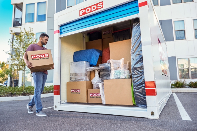 A man moving to an apartment with PODS carries a moving box toward a PODS container outside a modern apartment building.