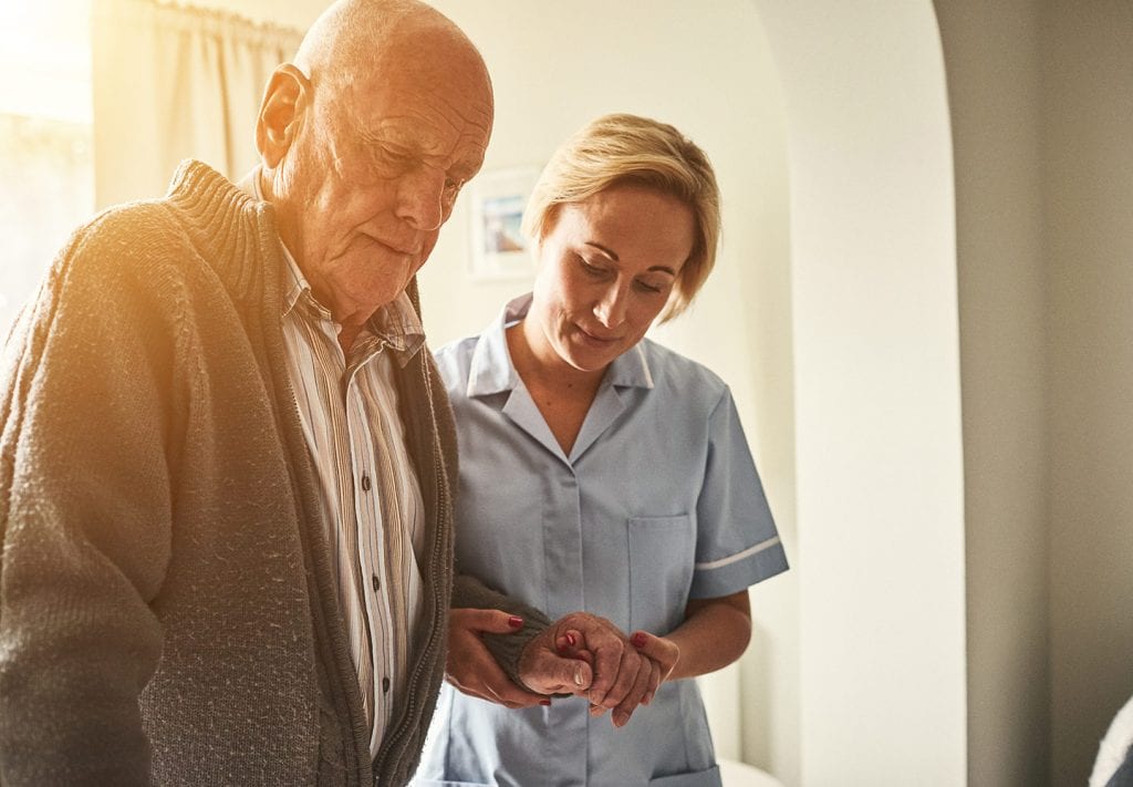 A woman helps an elderly man walk into the hallway in his home. 