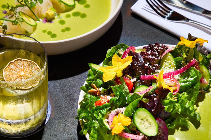  Expertly plated soup and salad from a San Francisco restaurant feature ingredients in vibrant hues. Off to the side sits a fancy cocktail with a dried lime slice floating on the top.