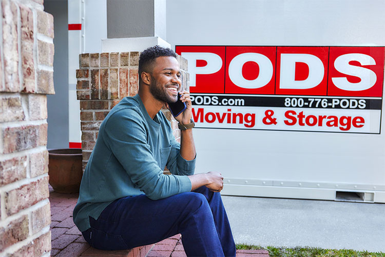 A man sits in front a PODS container in his driveway as he places a call on his cell phone