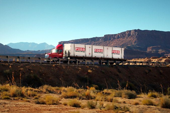 A PODS truck is transporting three PODS moving and storage containers cross-country across a rocky landscape.