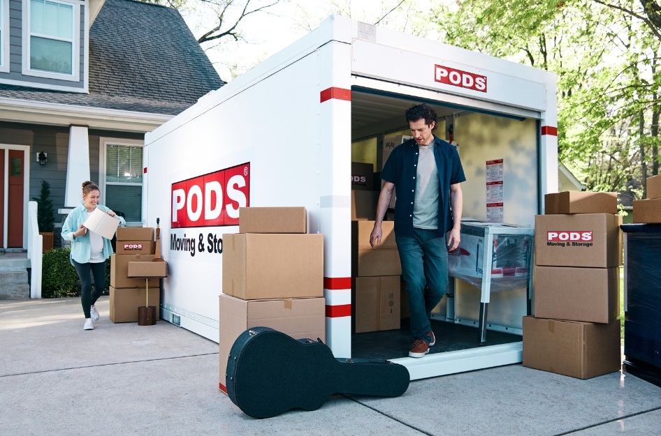 A man and woman are loading a PODS portable storage and moving container with furniture, moving boxes, and a guitar case.