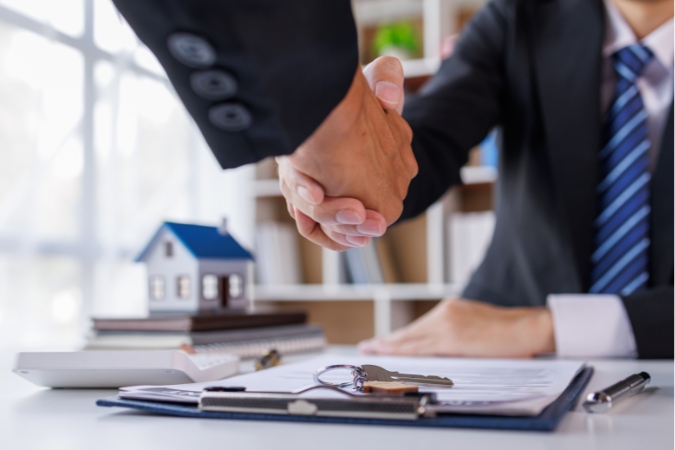 Closeup of a desk with a signed contract and set of keys on the surface and a man shaking hands with a seller after purchasing a new house.