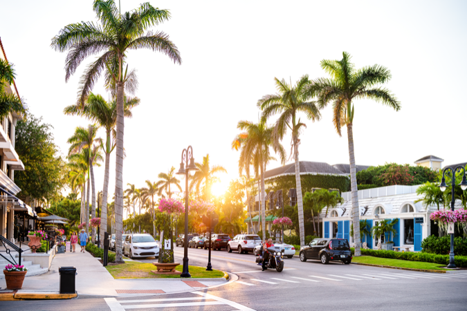 Sunset view of a palm tree-lined street in Naples — one of the safest cities in Florida — featuring cute storefronts and flowering plants hanging from quaint street lights.