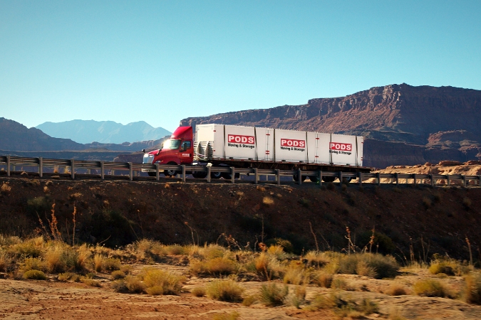 An 18-wheeler carrying PODS portable moving containers to Arizona, one of the best U.S. states for weather, drives along a desert highway with a view of mountains in the background.