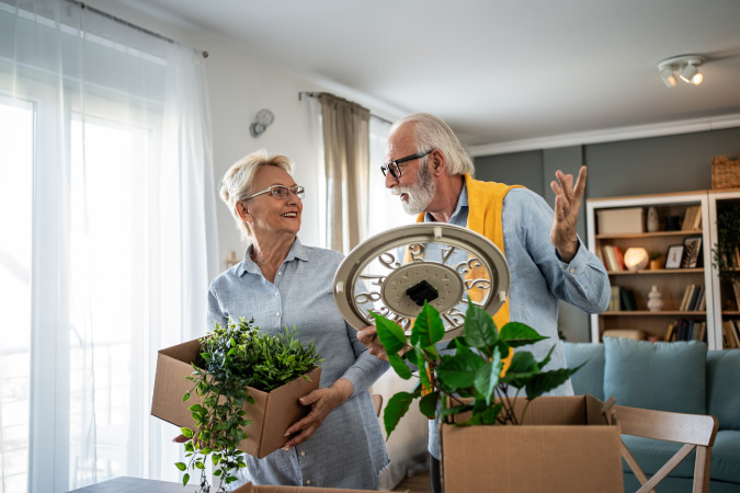 A retired couple is packing up moving boxes with houseplants and a wall clock.
