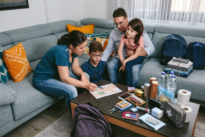 A family of four is sitting together in their living room, reviewing their family disaster preparedness plan, with various emergency items organized on the couch and table beside them.