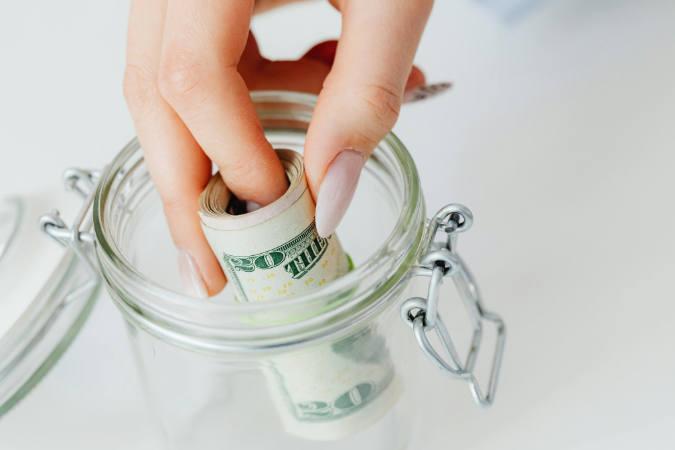 An image of a woman's hand putting a roll of money into a clear jar.