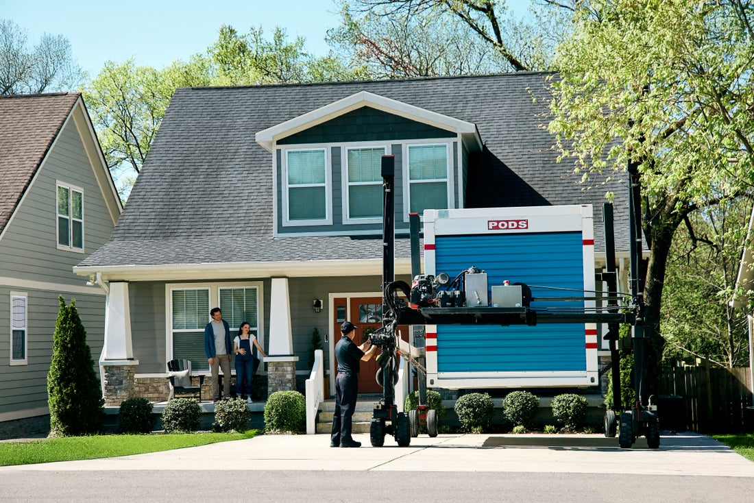 A PODS driver uses PODZILLA to position a PODS moving container in a residential driveway as the homeowners watch from the porch.
