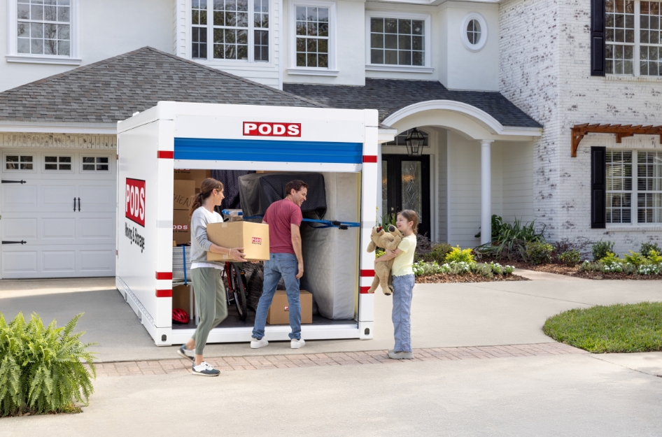 A family of three is unloading a PODS portable moving container in their driveway after moving to a more affordable city — one of the top reasons Americans are moving in 2026.