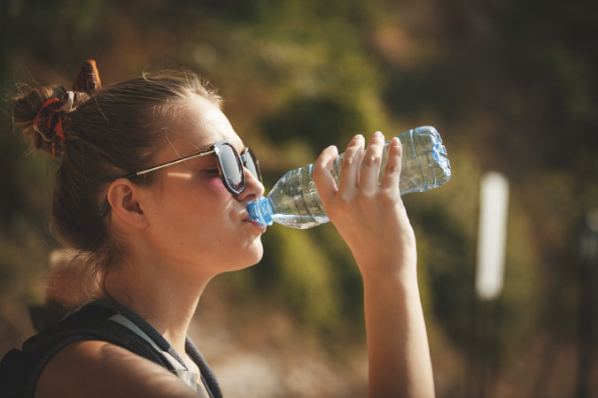 A young woman in sunglasses is drinking from a disposable water bottle on a hot, sunny day.