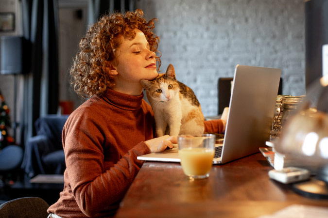 A young woman with curly hair is snuggling her cat as she uses her laptop to register the cat’s microchip online.