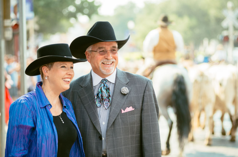 A happily retired couple laughs together as they pose for a photo. They’re both wearing black cowboy hats, and there’s a cattle drive happening in the background, though it’s out of focus.