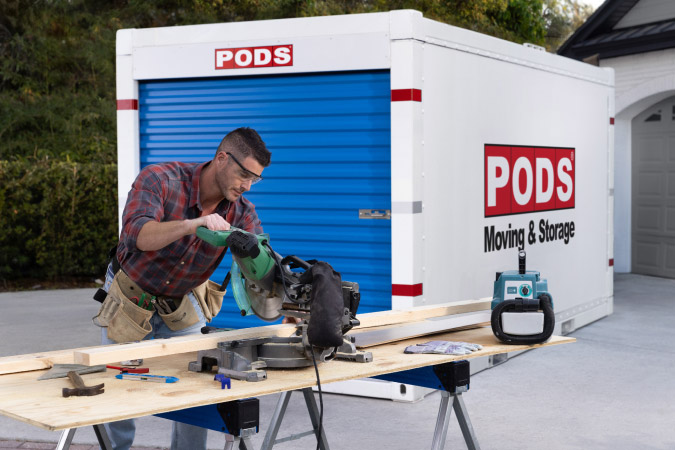 A man is remodeling after a natural disaster, using a mitre saw to cut a wooden board in his driveway beside a PODS portable storage container.