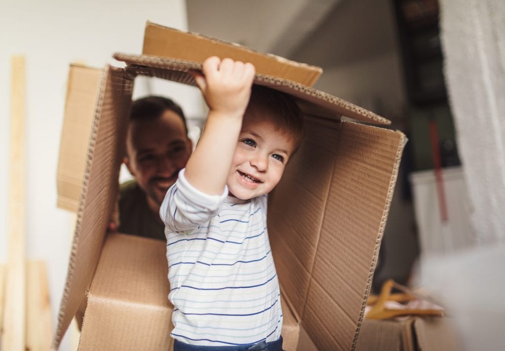 A little boy carries an empty, open cardboard. You can see his dad looking through the other end