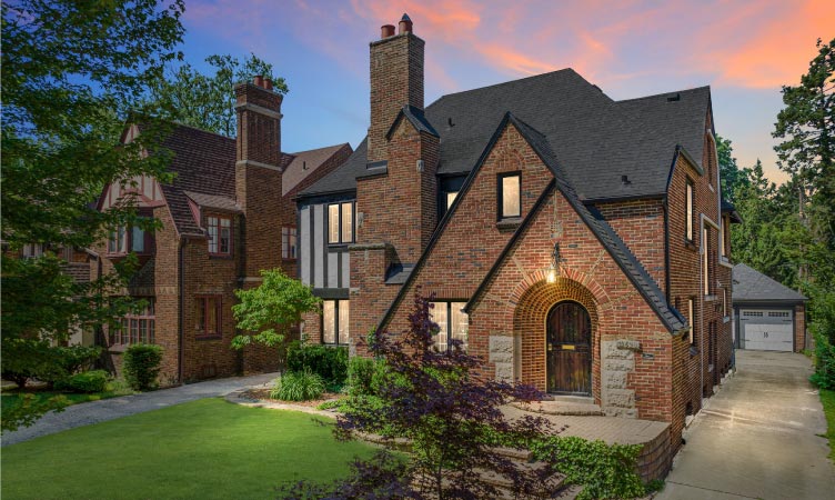 Upscale and historic brick homes in the Sherwood Forest neighborhood of Detroit. The outside light of the nearest home is illuminated as dusk begins to fall upon the neighborhood.