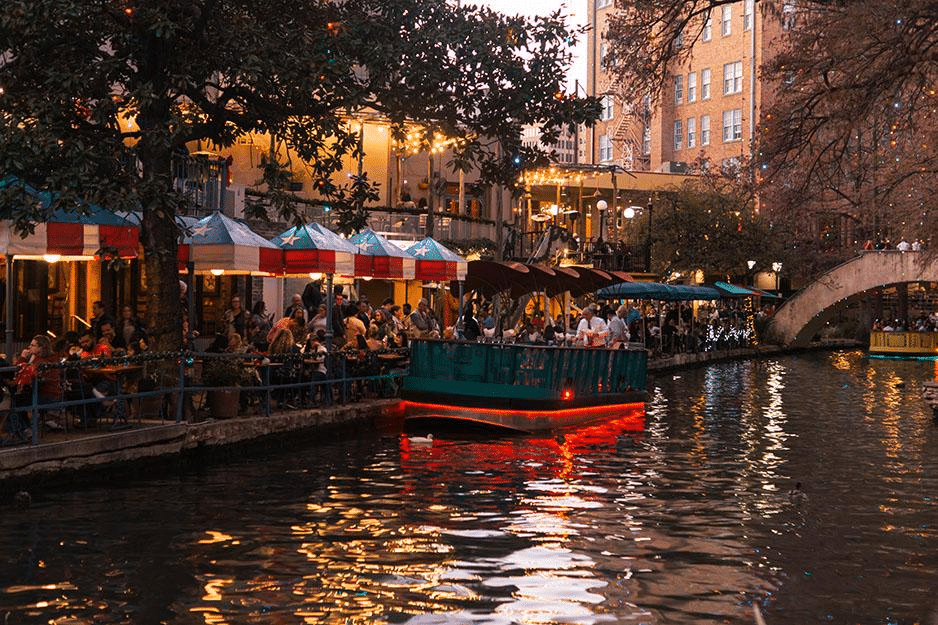 A small tour boat is floating down the San Antonio River as passersby dine and shop along the San Antonio River Walk.