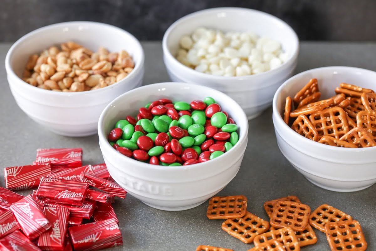 Bowls of peanuts, white chocolate chips, candy, and pretzels sit on a table. They’re ingredients for a Christmas snack mix, an easy-to-make, last-minute gift idea.