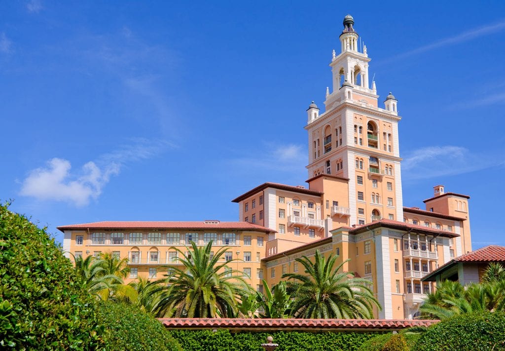 A beautiful building in Coral Gables, Florida. It's painted white and peach and there are palm trees in the foreground.