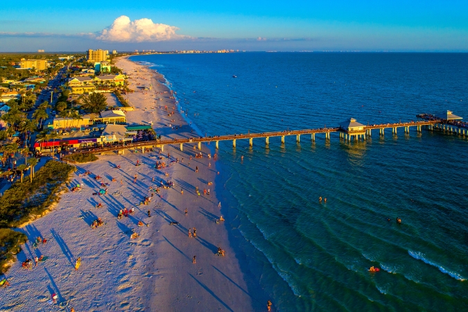 Aerial sunset view of Fort Myers Beach featuring the pier, scattered beachgoers along the shoreline, calm waves, and resort buildings in the background.