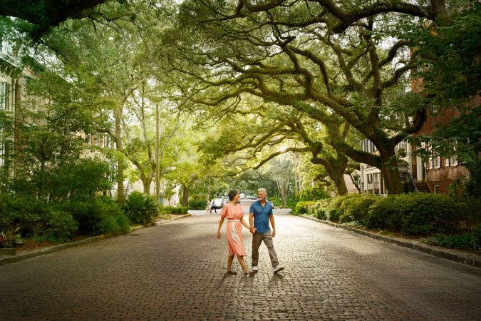 A retired couple is crossing a shady cobblestone street in Savannah, Georgia.