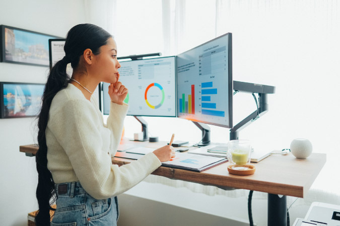 A woman is standing at her desk working on two side-by-side monitors, which are part of her home office setup.